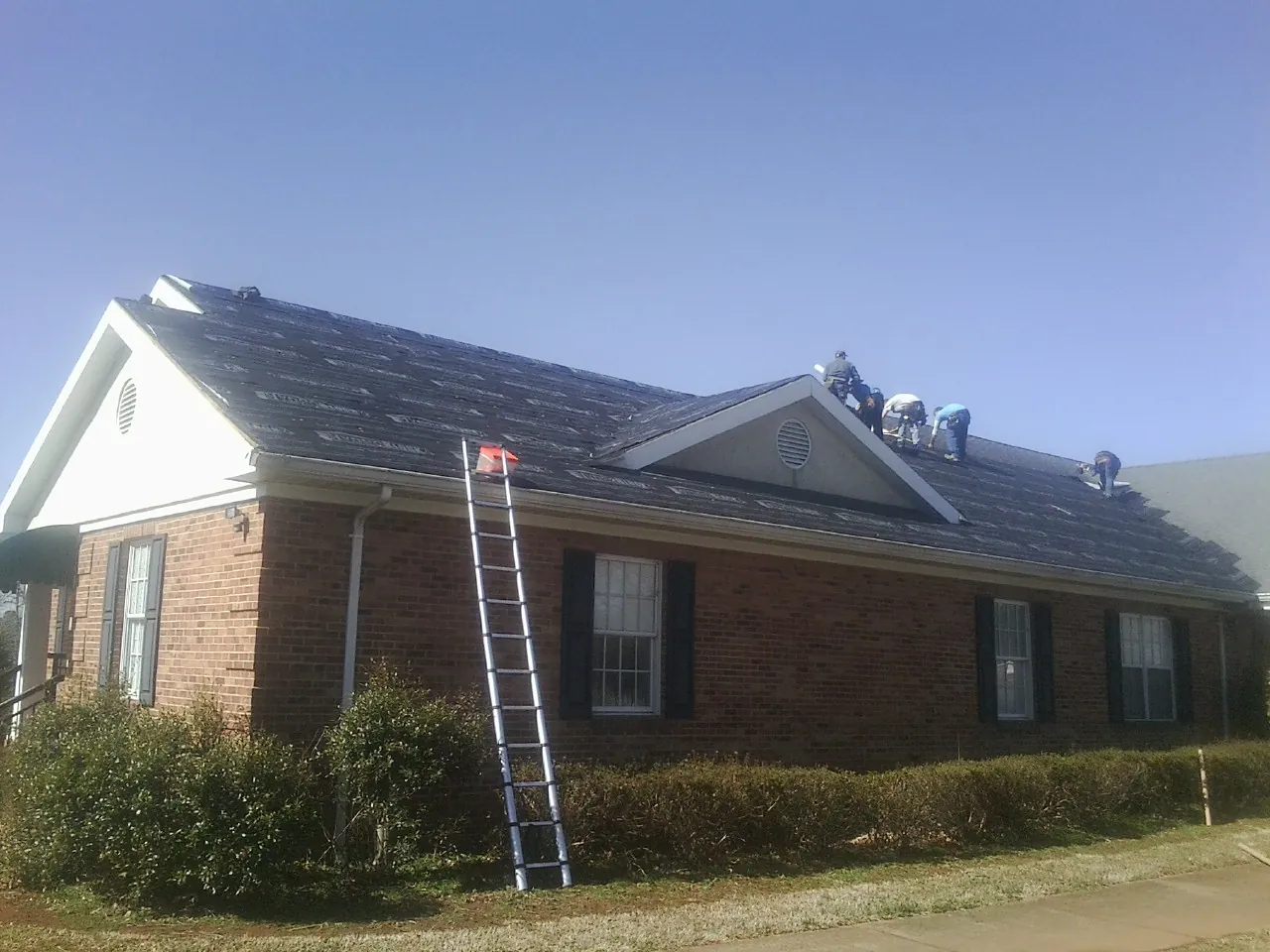 Crew tearing shingles off a brick ranch roof with a ladder set — Metal Roof Repair work by our crew in Parshall