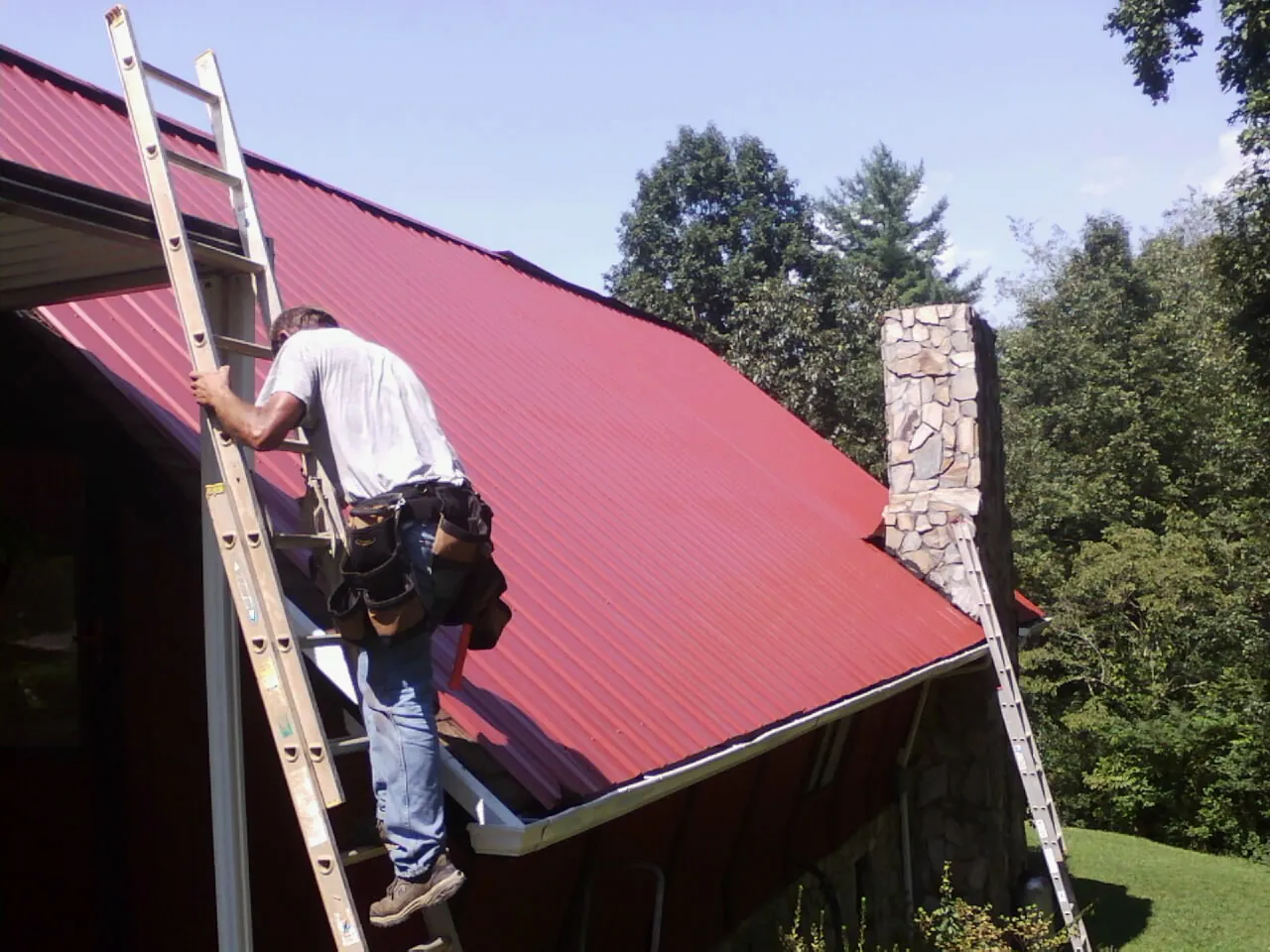 Roofer on a ladder beside a red metal roof and stone chimney — Soffit & Fascia Repair by Roofing Brothers in Parshall